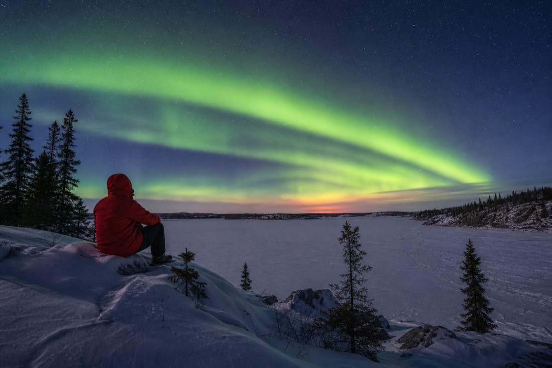 Traveler in a red parka watching a bright aurora band over a frozen lake outside Yellowknife