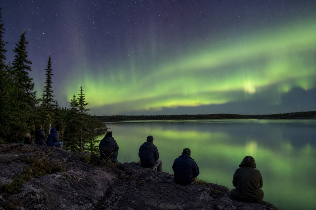 Summer crowd on a rocky lakeshore in Yellowknife with spruce trees and bright water behind them