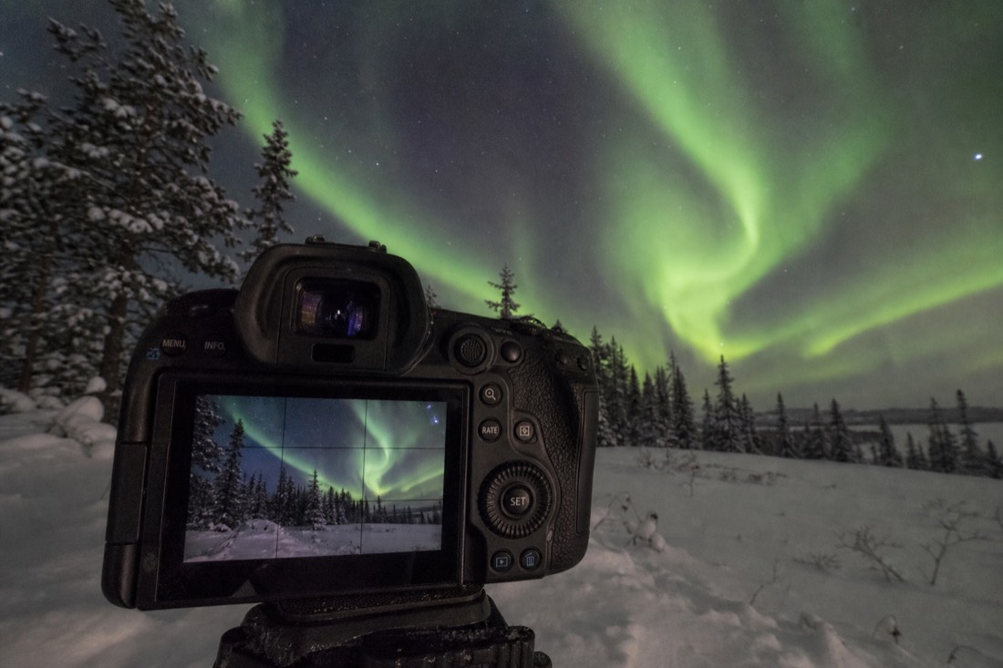 Camera screen showing an aurora exposure preview while the real green curtains fill the sky behind it