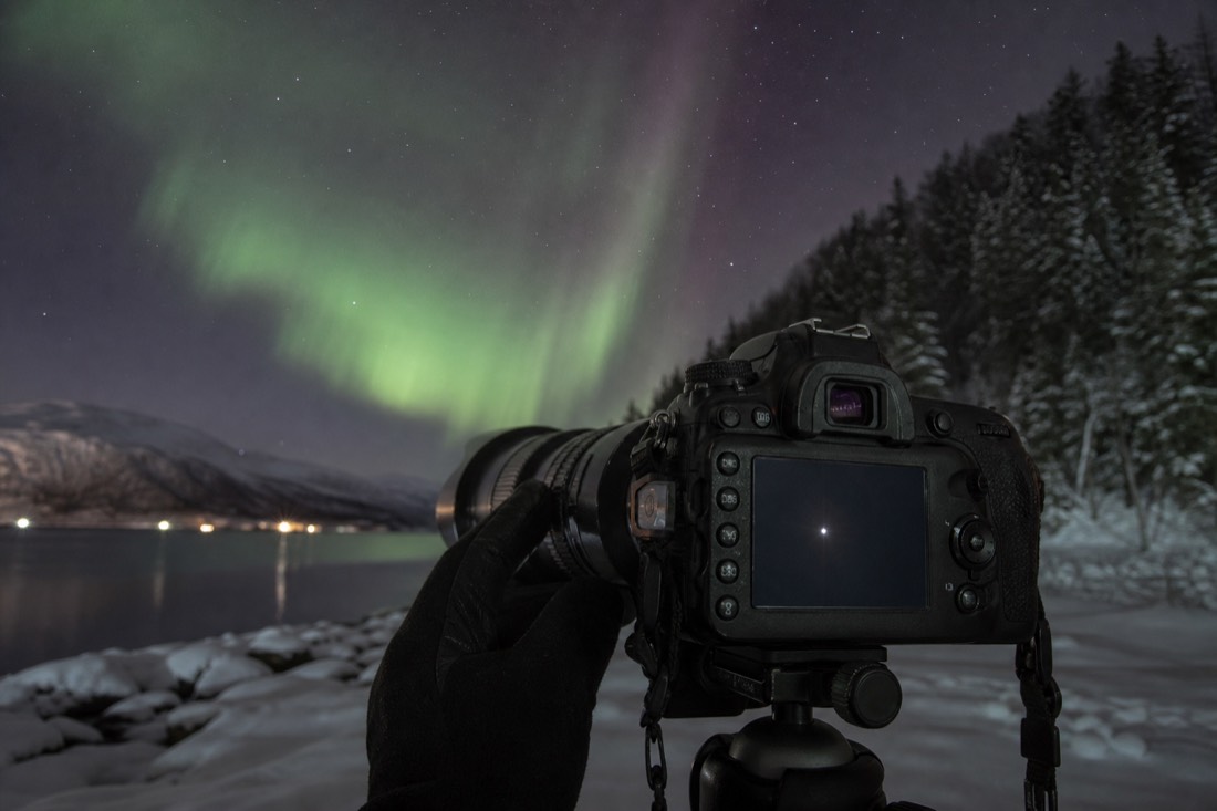 Gloved hand turning a lens focus ring while the camera's magnified live view shows a single bright star