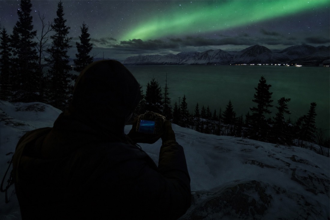 Photographer reviewing a too-dark aurora frame on the back of a camera at a dark lakeside viewpoint