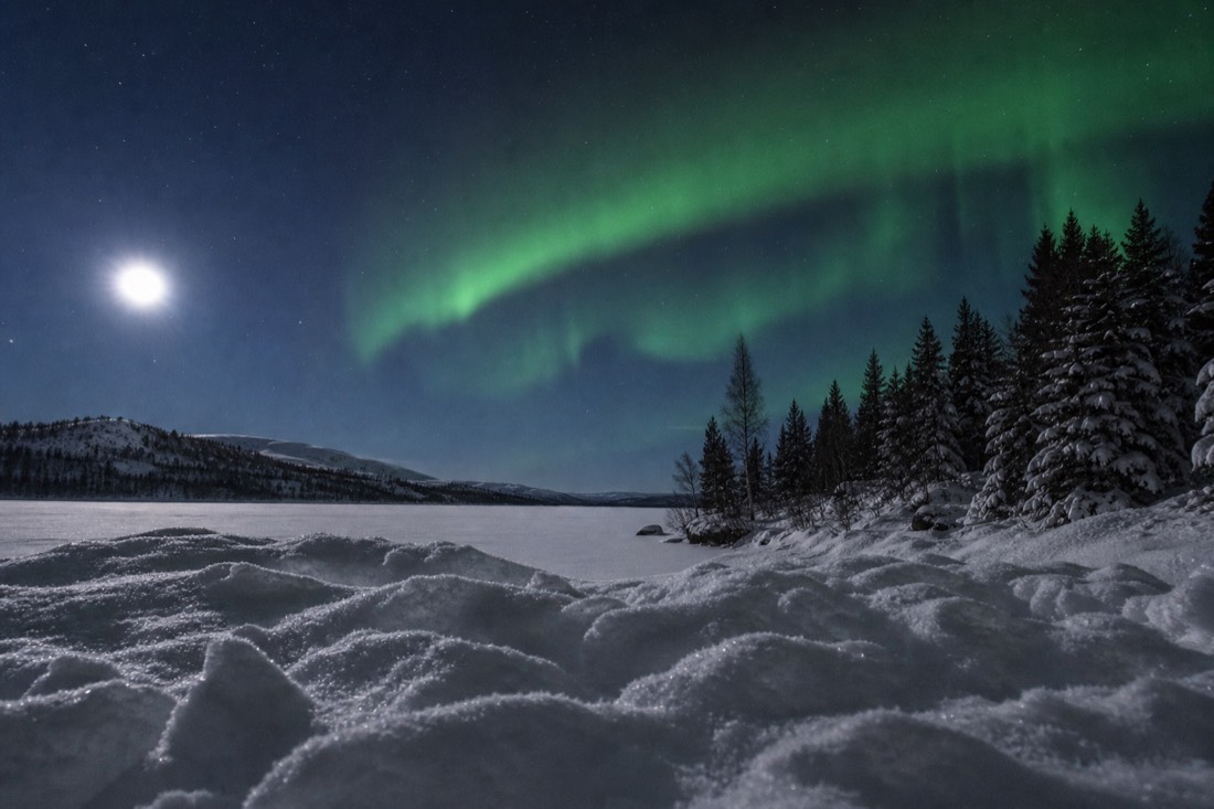 Aurora arc over a moonlit snow-covered lake with bright snow texture visible in the foreground