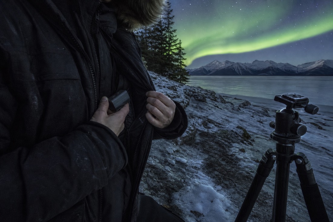 Photographer sliding a spare camera battery into an inner parka pocket beside a tripod on frozen ground
