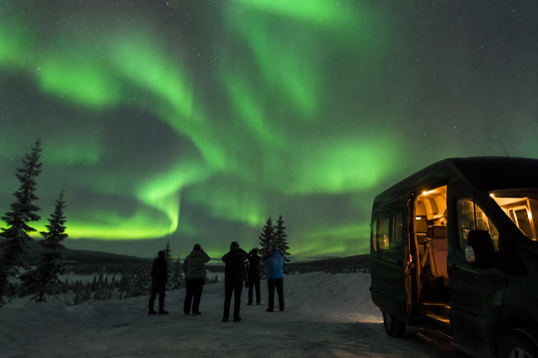 Tour photographer framing guests under bright aurora beside a heated van at a dark viewpoint