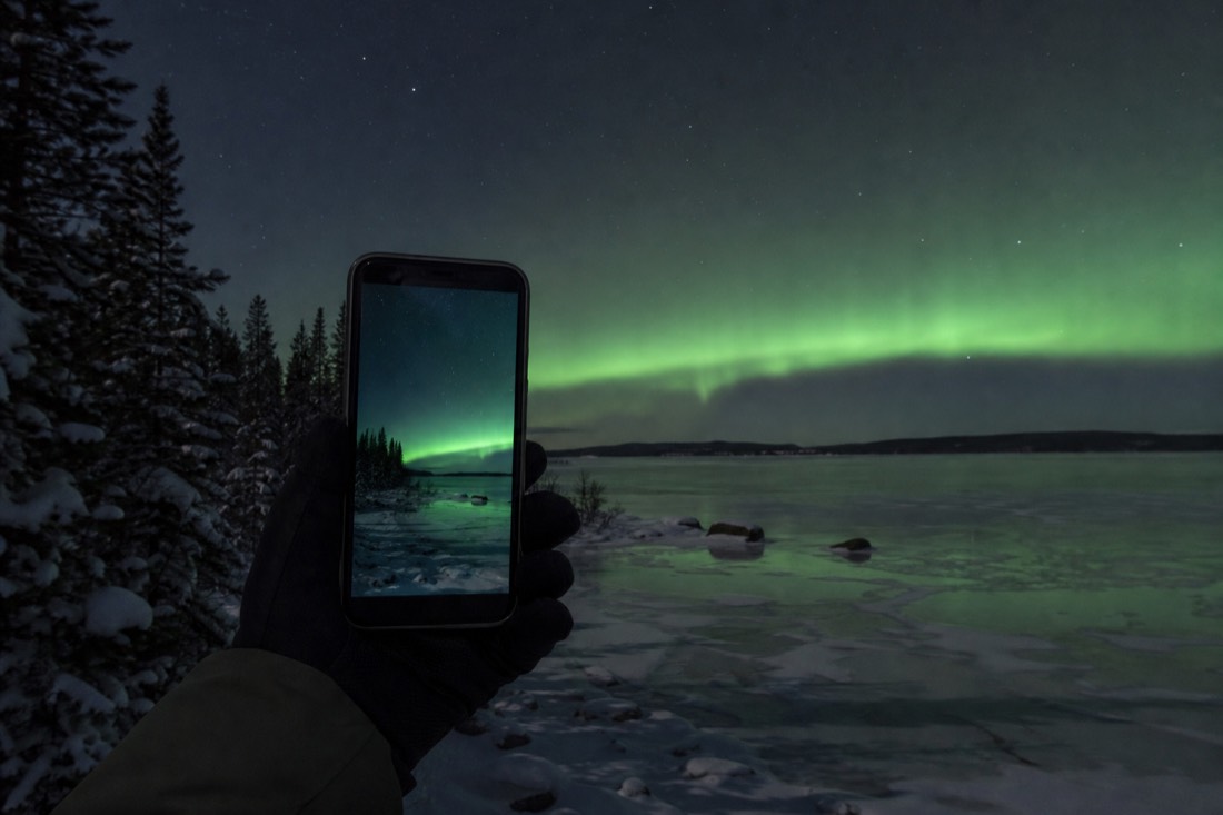 Phone screen showing a green aurora band while the same lights glow faintly over a dark frozen lake behind it