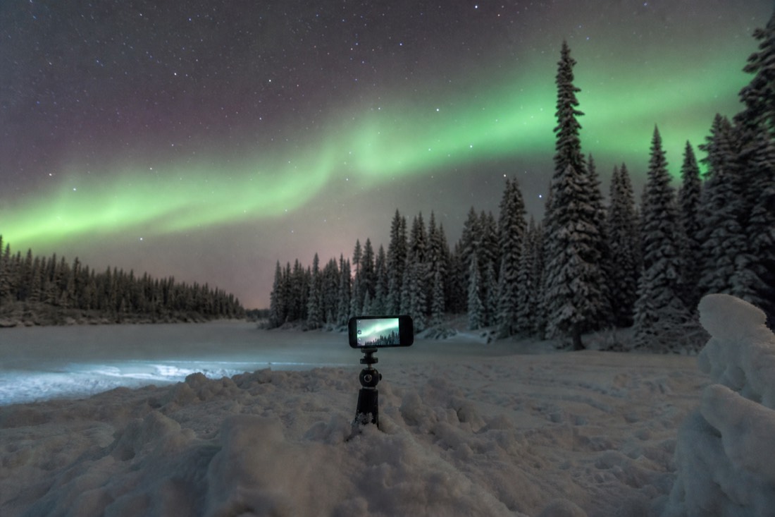 Phone wedged into packed snow on a mini tripod, screen glowing, aurora arc overhead