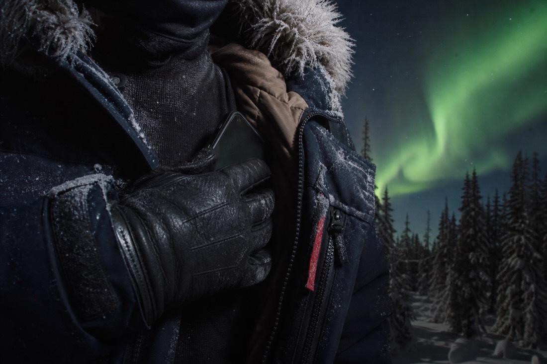 Gloved hand tucking a phone inside a parka against a base layer with frost on the parka fur ruff