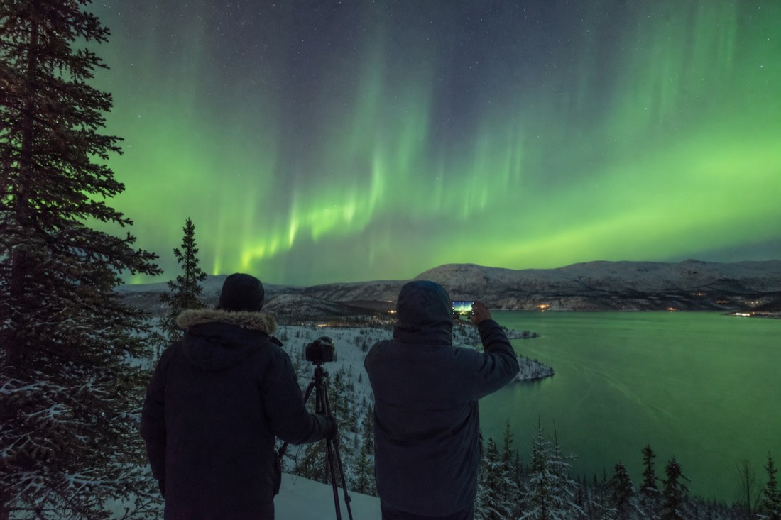 Guide with a tripod-mounted camera beside a visitor shooting the same aurora on a phone at a lakeside viewpoint
