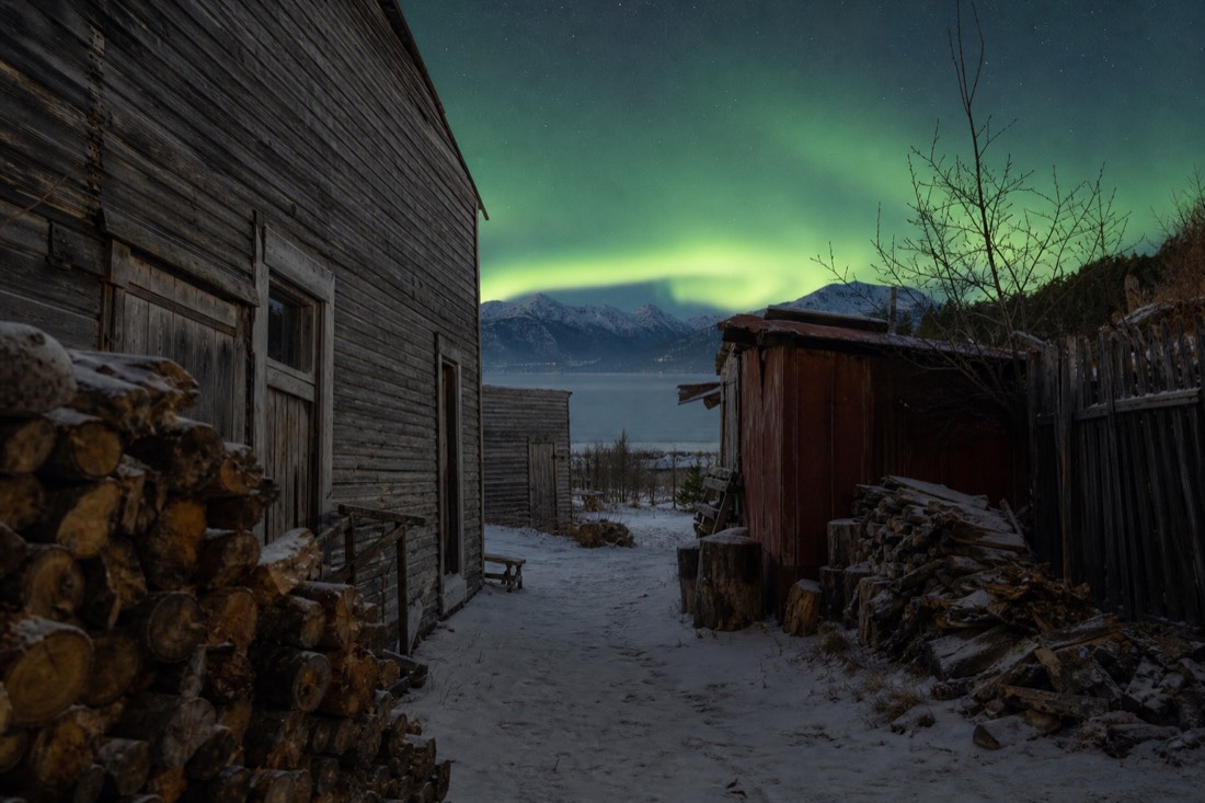 Narrow Old Town lane with gold-rush era shacks, stacked firewood and the frozen bay visible at the end