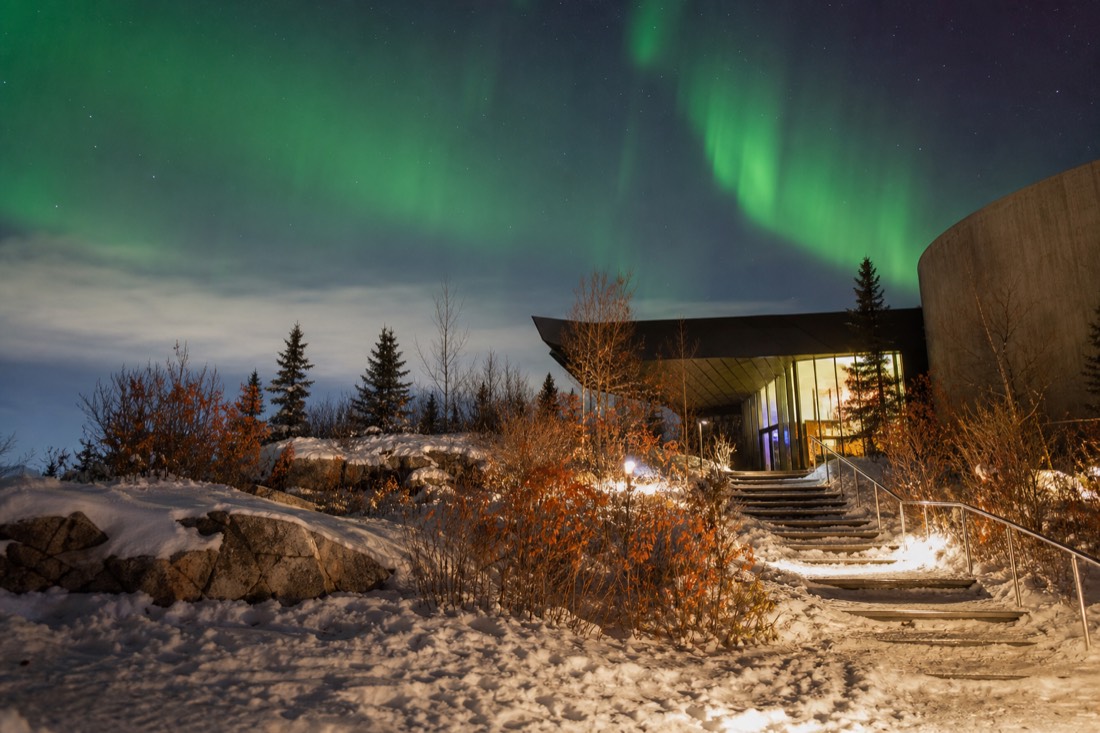 Museum entrance framed by autumn shrubs and Canadian Shield rock beside Frame Lake in Yellowknife