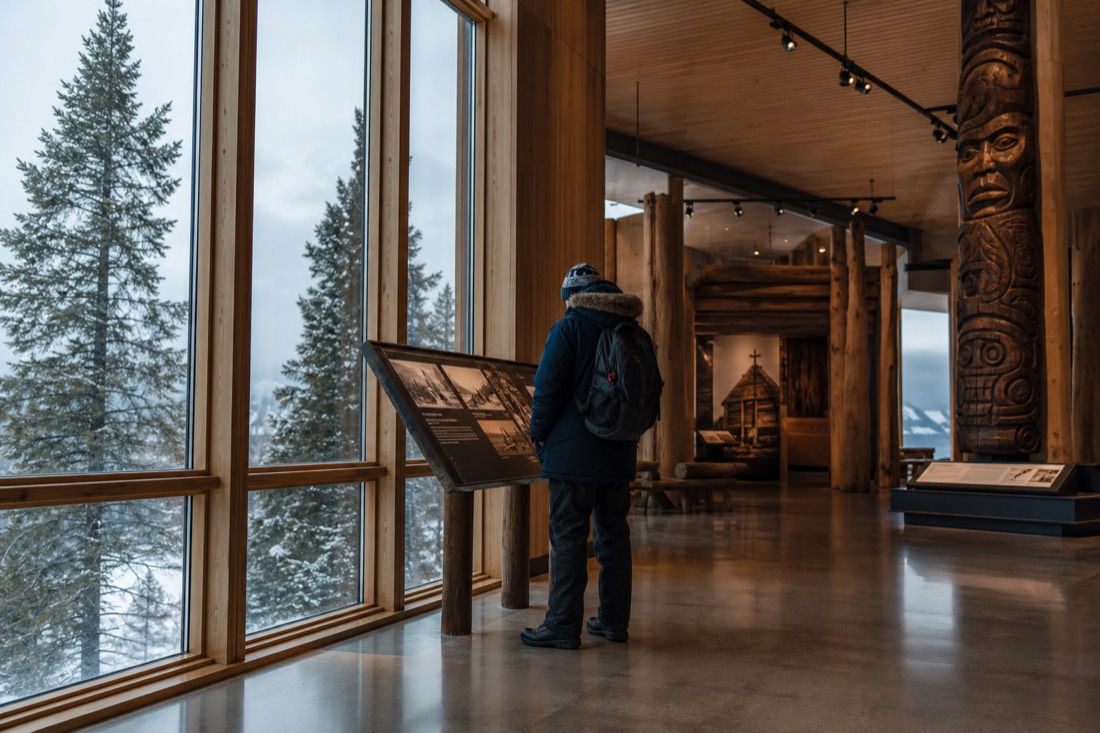 Visitor in winter layers reading an exhibit panel in a bright museum atrium with snow visible outside