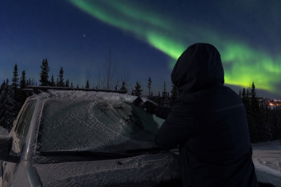 Driver scraping heavy frost from a car windscreen in the blue half-light of a Yellowknife winter morning