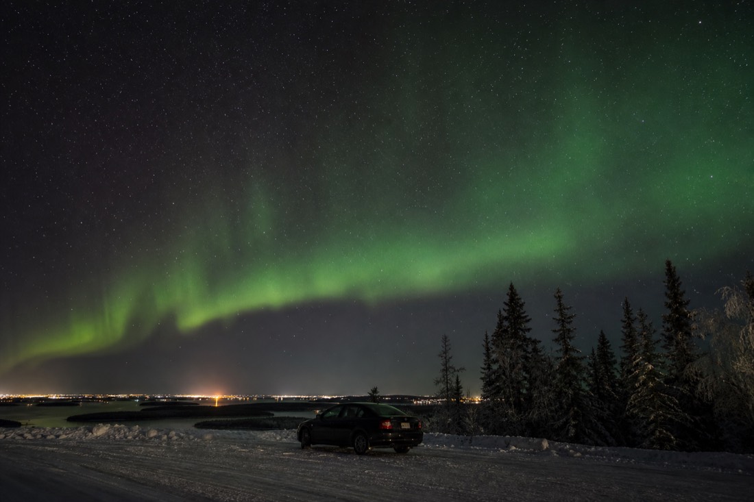 Lone parked car at a dark roadside pull-out under a green aurora arc outside Yellowknife