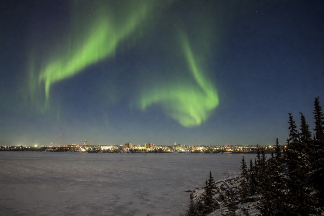 Green aurora over the small lit skyline of Yellowknife with frozen Great Slave Lake in the foreground