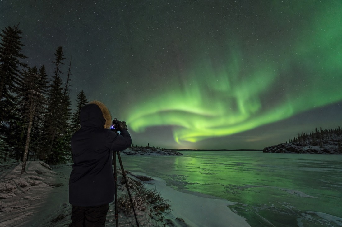 Traveler in a heavy parka photographing bright green northern lights over a frozen lake near Yellowknife