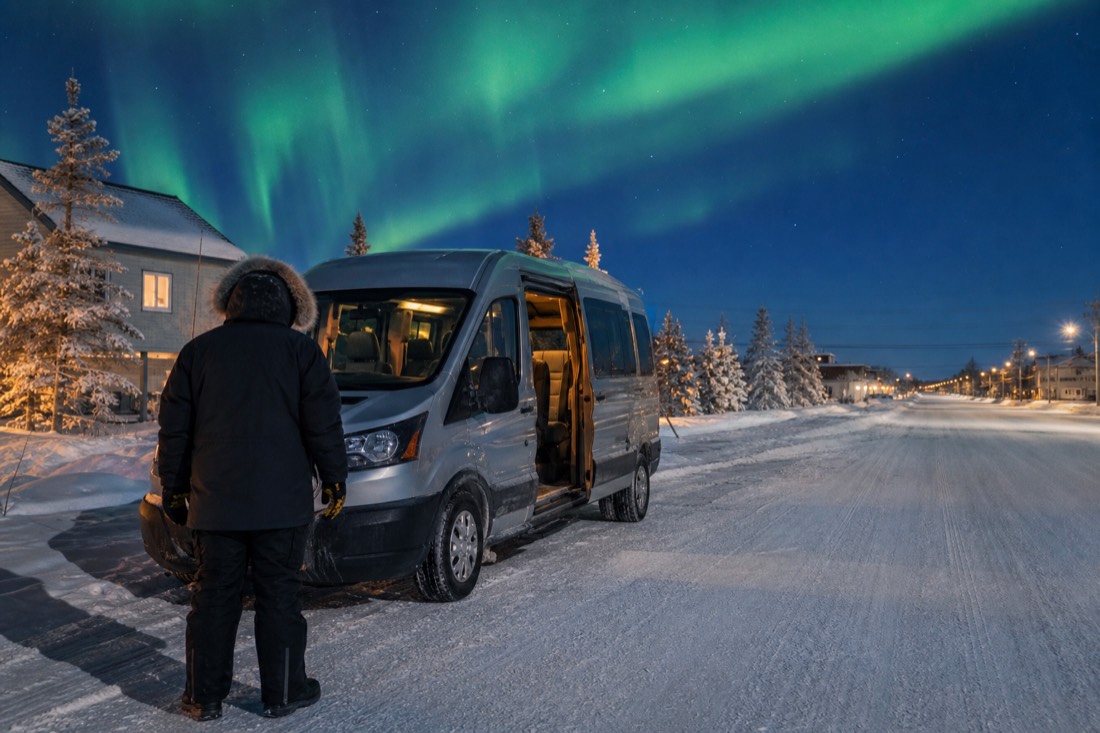 Guide in a heavy parka standing beside a warm tour van on a snowy Yellowknife street at blue hour before an evening aurora pickup
