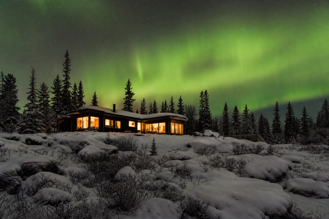 Heated aurora lodge windows glowing warm under a green sky in the forest outside Yellowknife