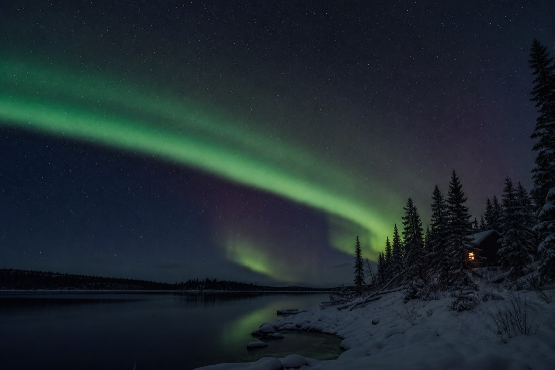 Aurora arch above a remote dark lakeshore with one lit cabin window and no town glow on the horizon