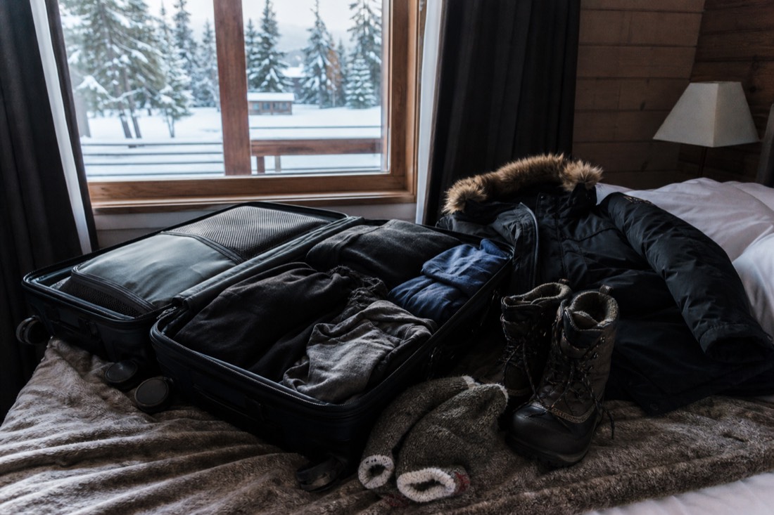 Open suitcase on a hotel bed with thermal base layers and wool socks beside a bulky rented parka and boots