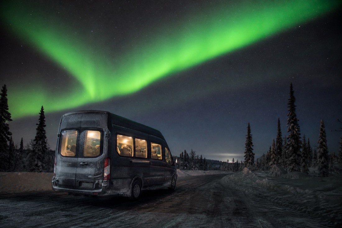 A heated tour van parked on a dark snow-packed road outside town under a bright green aurora band