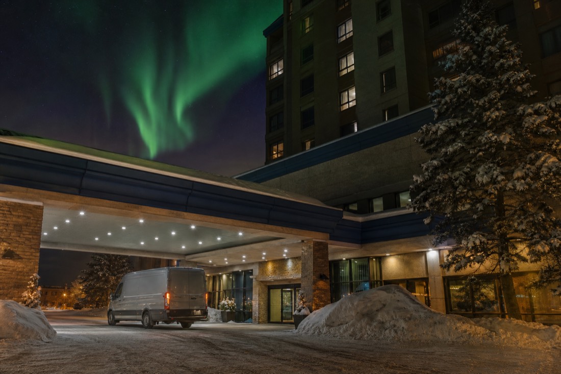 A large downtown Yellowknife hotel entrance at night with snow piled by the doors and a van waiting under the canopy
