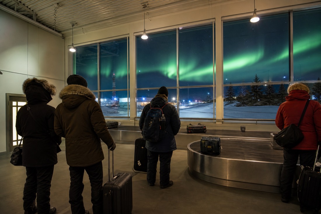 Compact northern airport terminal interior with passengers in parkas collecting bags near a small baggage carousel