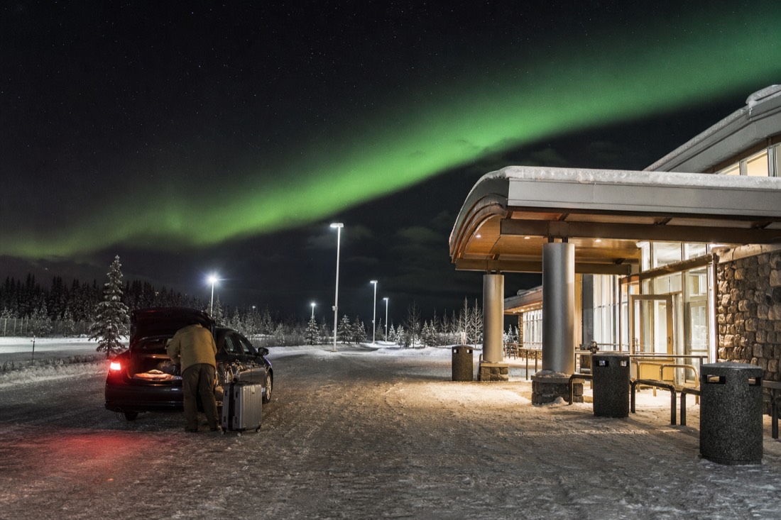 Traveller loading luggage into a waiting taxi outside a small snowy airport terminal under winter floodlights