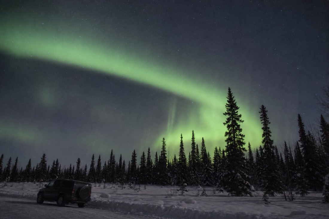 Green aurora arc over a dark spruce treeline with a single vehicle parked on a snowy pull-off