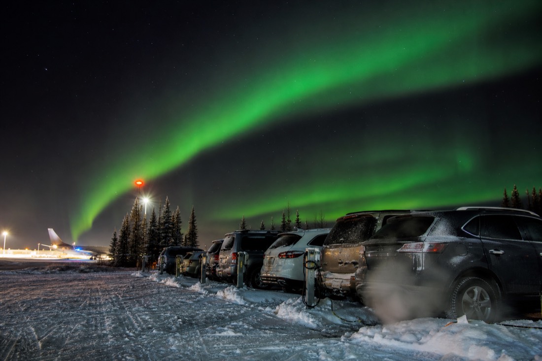 Cars parked in a snowy airport lot with block heater cords plugged into posts and exhaust vapour in the cold air