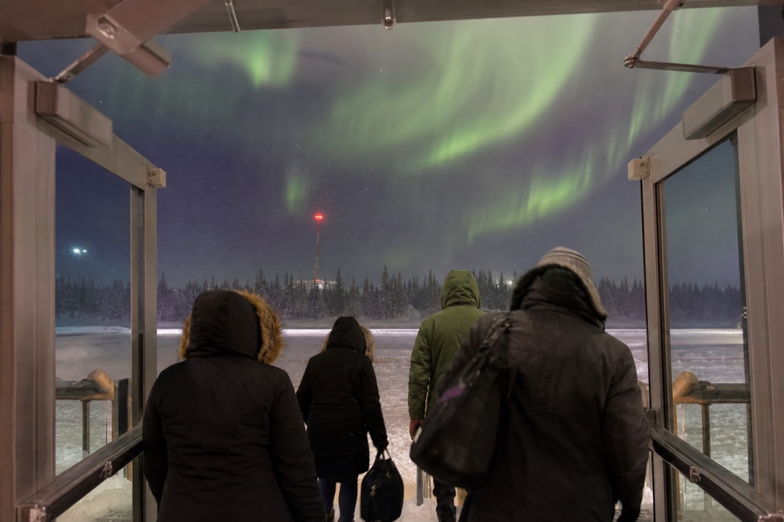 Arriving passengers pulling parkas and hats on inside an airport vestibule before stepping out into blowing snow at night