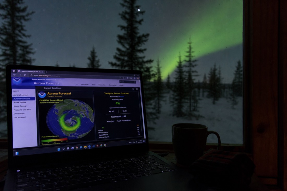 Aurora forecast dashboard glowing on a laptop screen beside a window looking out on a dark northern night
