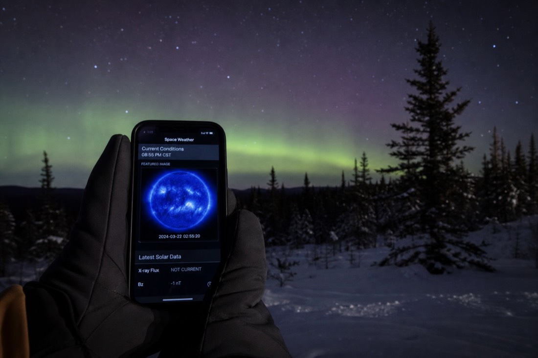Gloved hands holding a phone showing a space weather page with a dark treeline and stars behind
