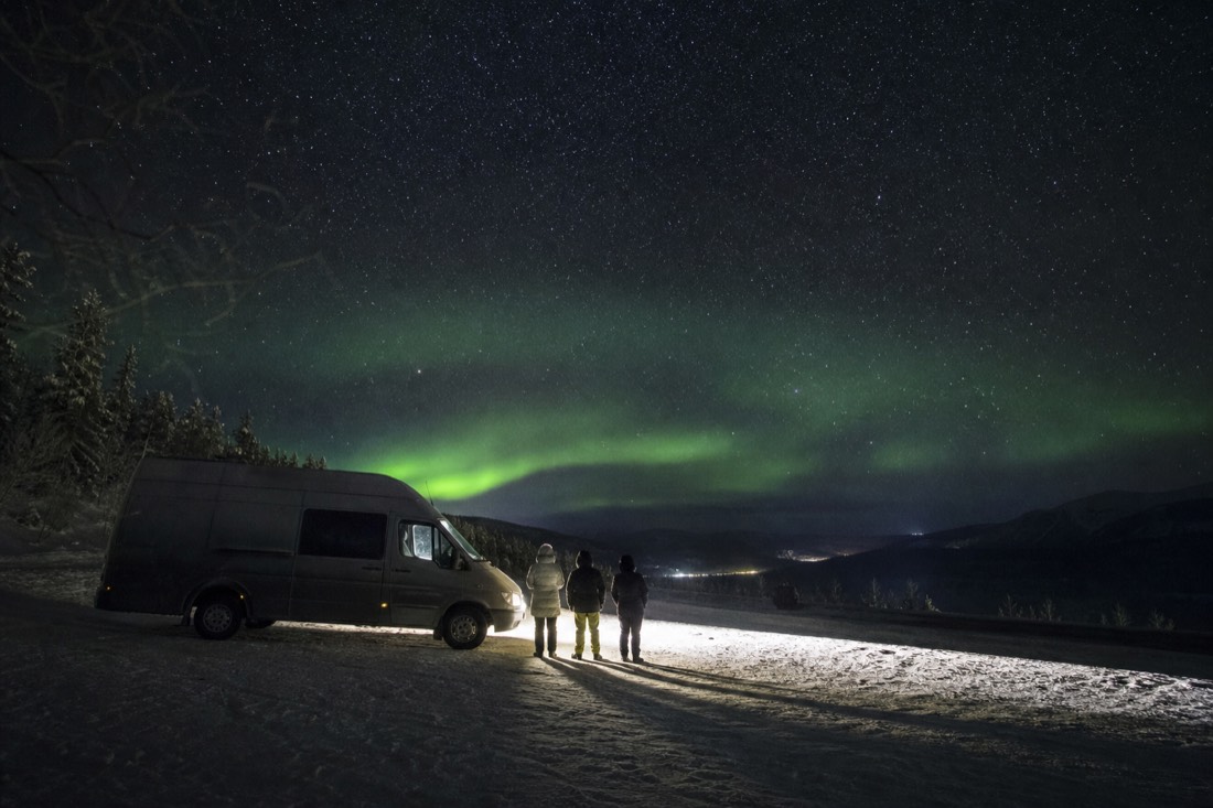 Van parked on a snowy pullout under a clear starry sky with guests standing in the headlights' glow
