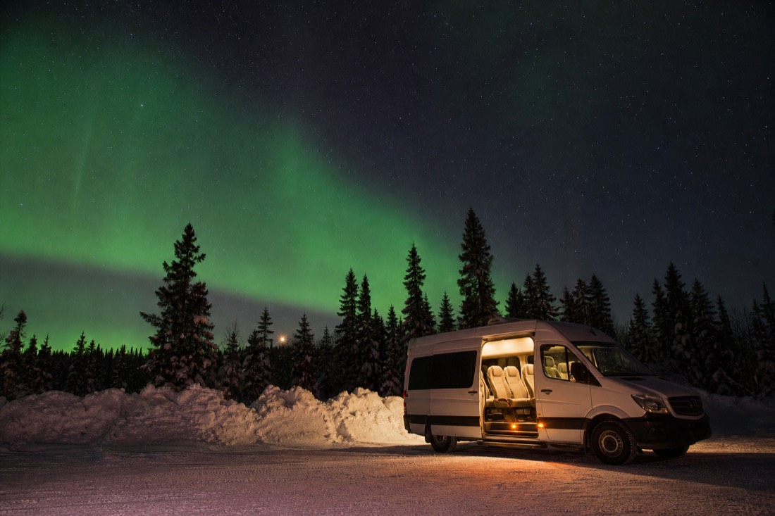 Aurora tour van with its interior light on parked beside a snowbank near midnight while faint green lights rise behind the trees