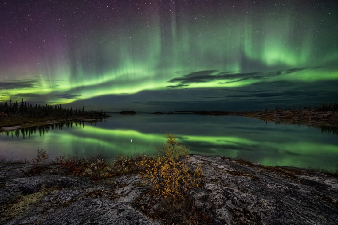 September aurora reflected in the open water of a lake near Yellowknife with bare rock and autumn scrub in the foreground