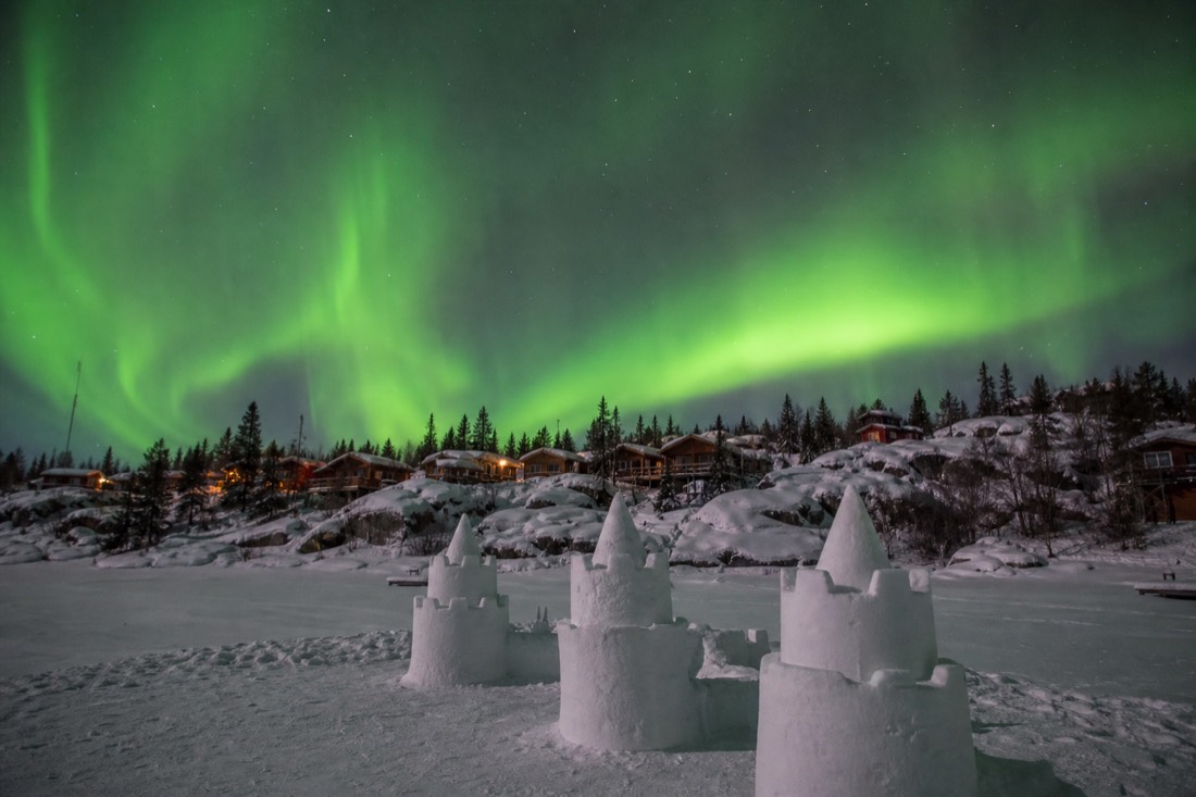 Snow castle towers rising from the frozen surface of Yellowknife Bay with Old Town cabins on the shore behind