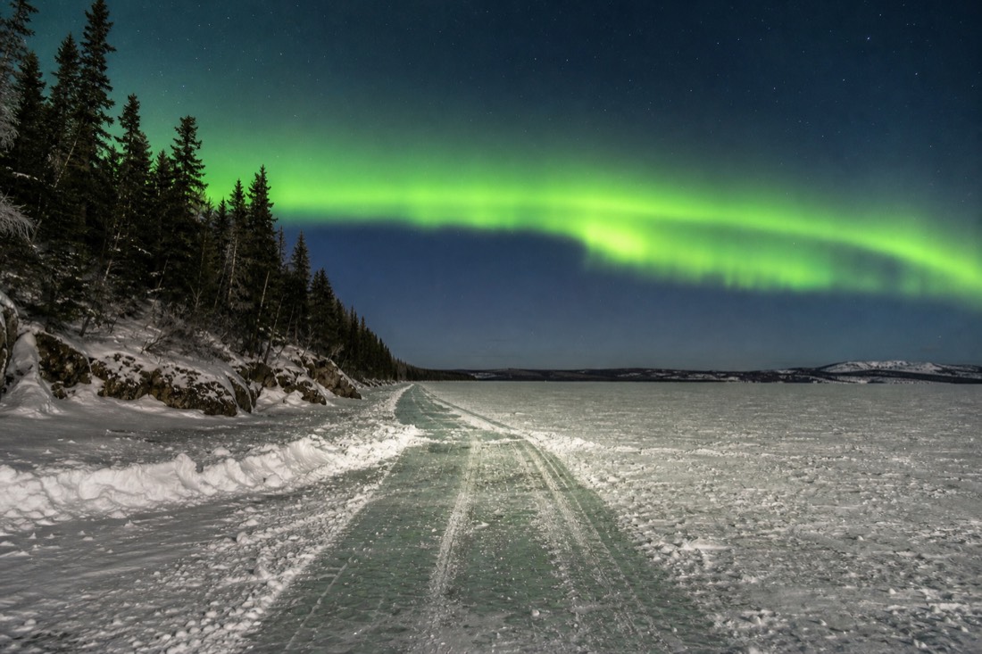 Green aurora arc above a frozen lake with a plowed ice road line running toward the horizon