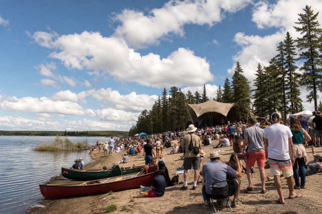 Crowd on a sunny lakeshore at an outdoor summer music festival with canoes pulled up on the sand