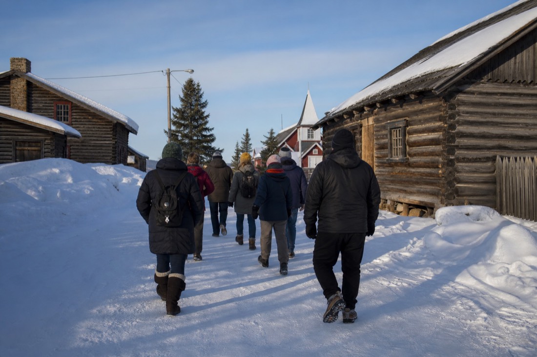 Small tour group walking past historic log buildings in Old Town Yellowknife on a bright winter afternoon