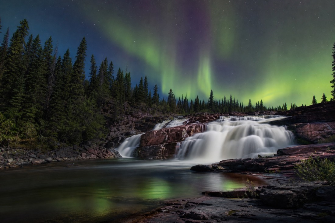 Waterfall pouring over pink granite ledges into a river framed by spruce forest in full summer
