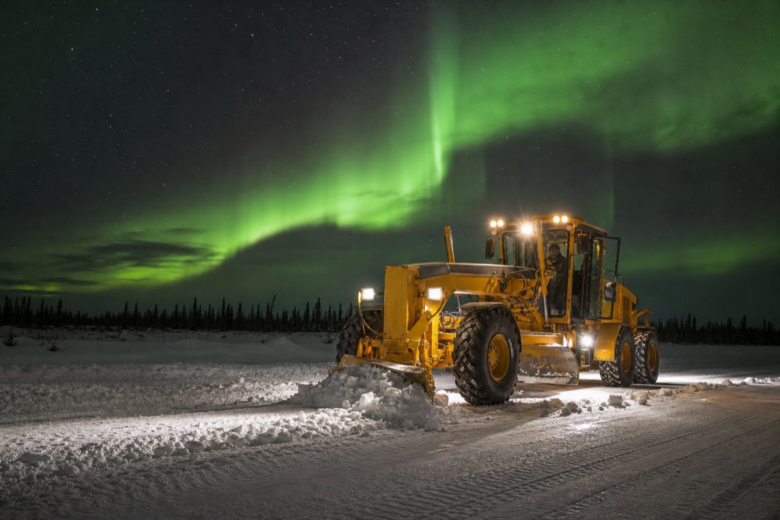 Road grader clearing snow from a wide ice road surface with spruce shoreline in the far distance