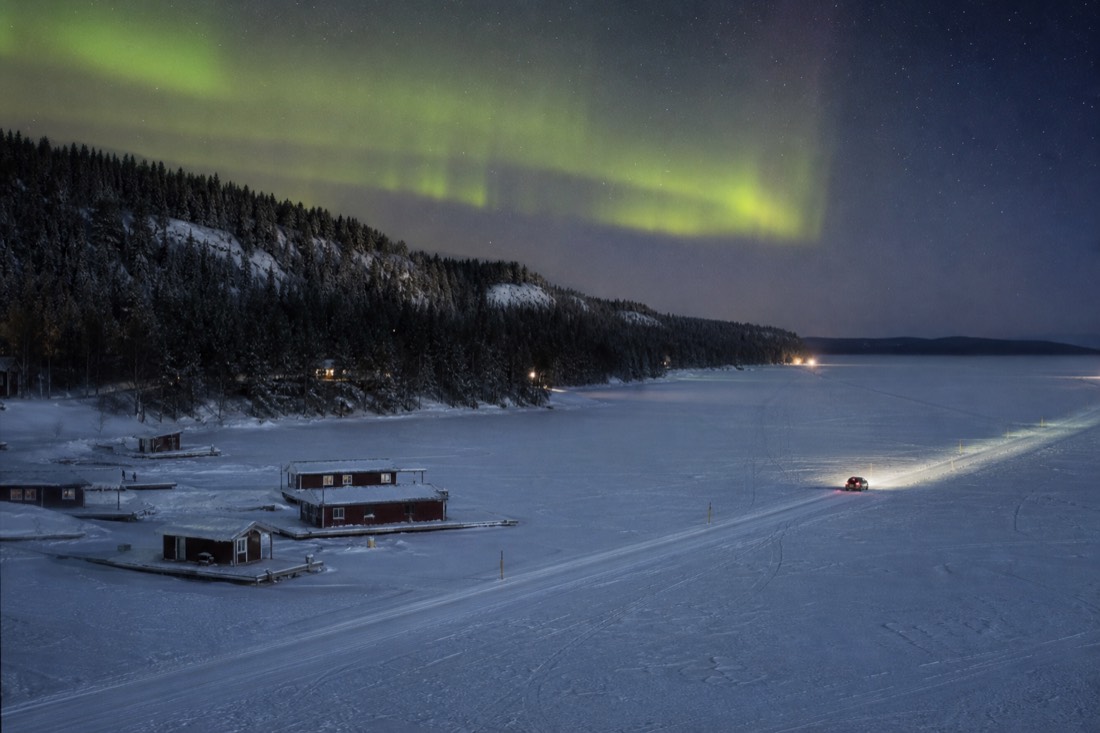 Small car driving the marked ice road across a frozen bay with houseboats frozen in near the shoreline