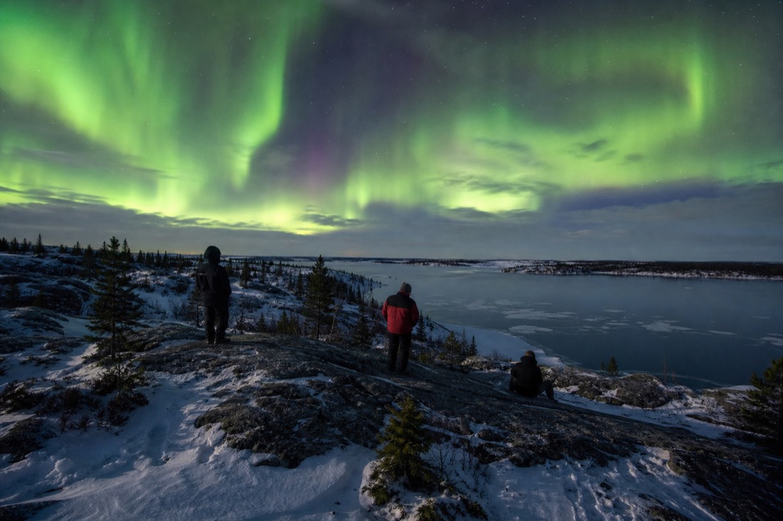 Small group of visitors on a rocky Yellowknife lookout in bright late April evening light with open water starting to show