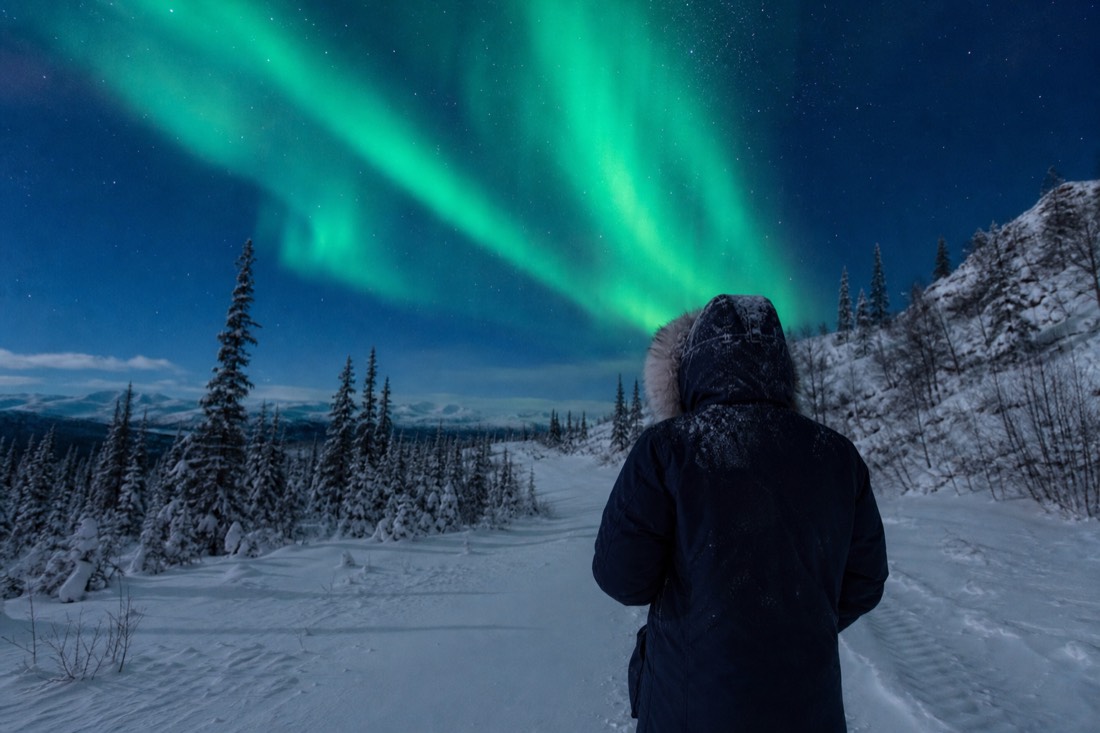 Traveler in a heavy rented parka with a frosted fur hood standing on a snowy trail in flat February light