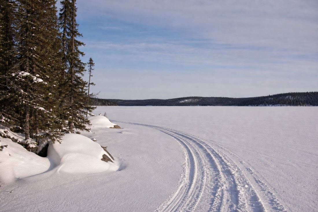 Wide sunlit February view across a frozen lake with spruce treeline and a snowmobile track in the foreground