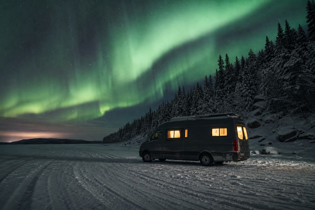 Bright green aurora curtain arching over a snow-covered frozen lake with a heated tour van parked on the ice