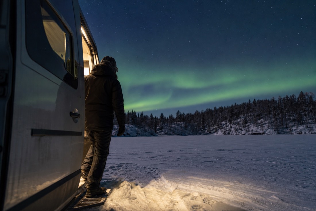 Traveler in full winter gear stepping out of a heated van onto a frozen lake under a faint green January aurora