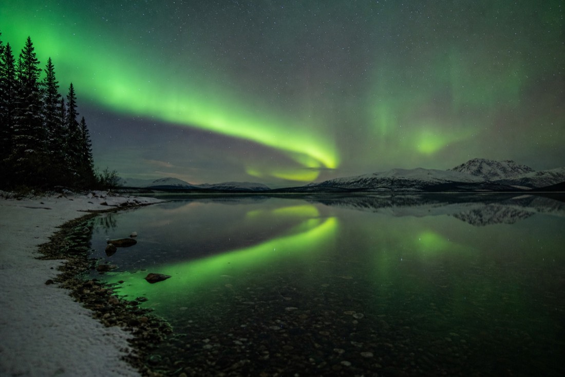 Green aurora arc reflected in unfrozen lake water with a thin line of snow on the shore