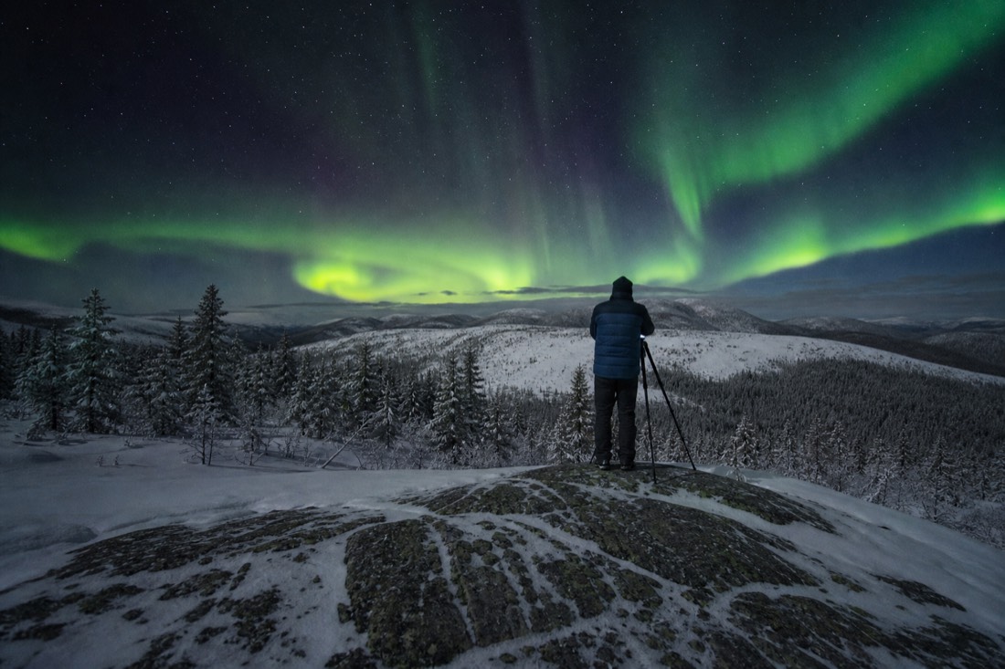 Photographer with a tripod standing on bare rock at night in a light jacket rather than a parka