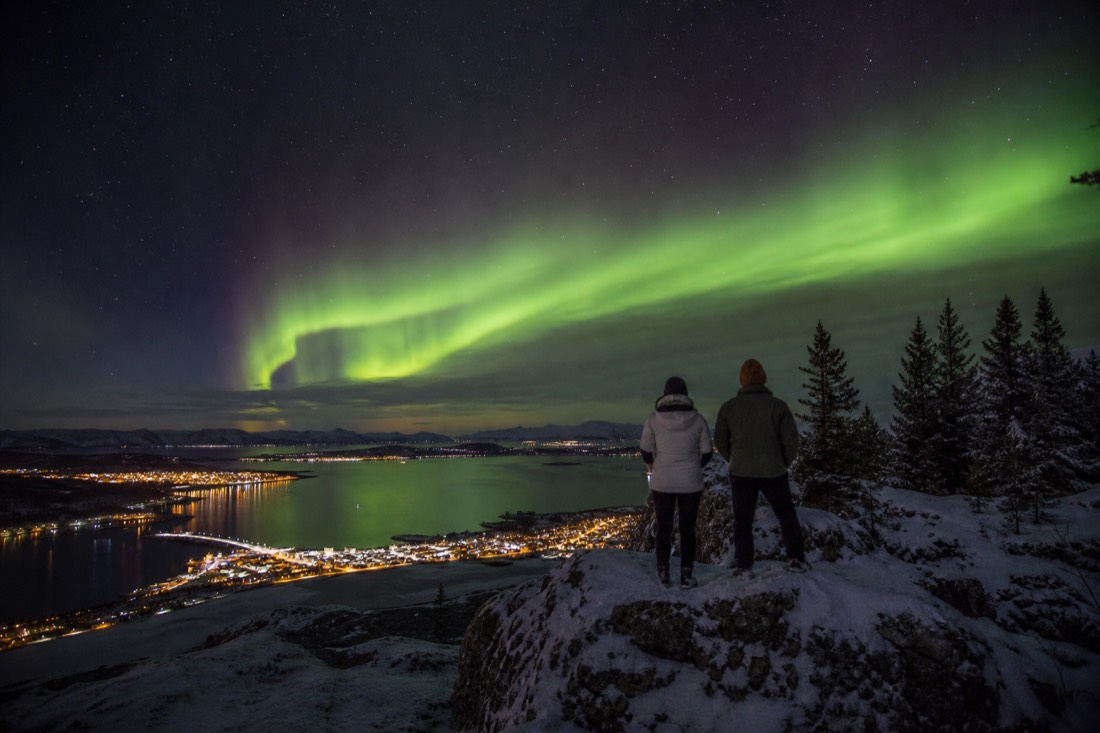 Two travellers in light winter jackets looking at a dark northern sky from a rocky lookout above town